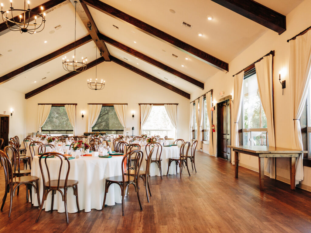 A spacious event hall with wooden floors, round tables covered in white cloths, wooden chairs, chandeliers, large windows with cream curtains, and rustic décor set up for a gathering.