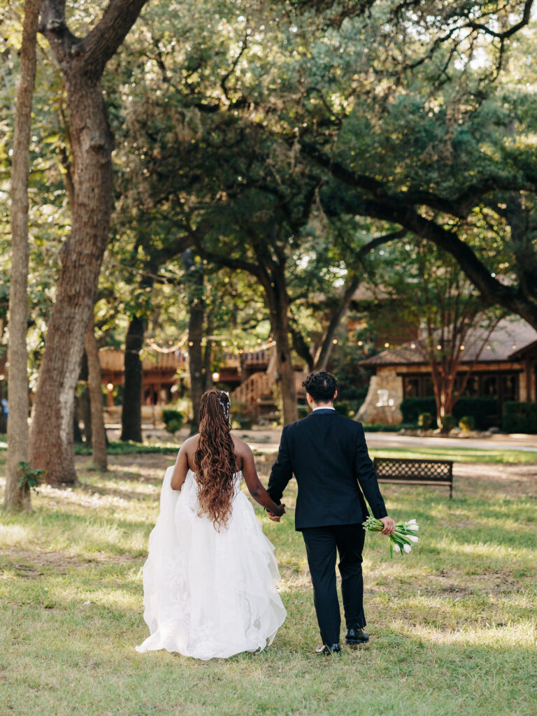 A bride in a white dress and a groom in a black suit walk hand in hand across a grassy area surrounded by tall trees, with the groom holding a bouquet and a wooden building visible in the background.