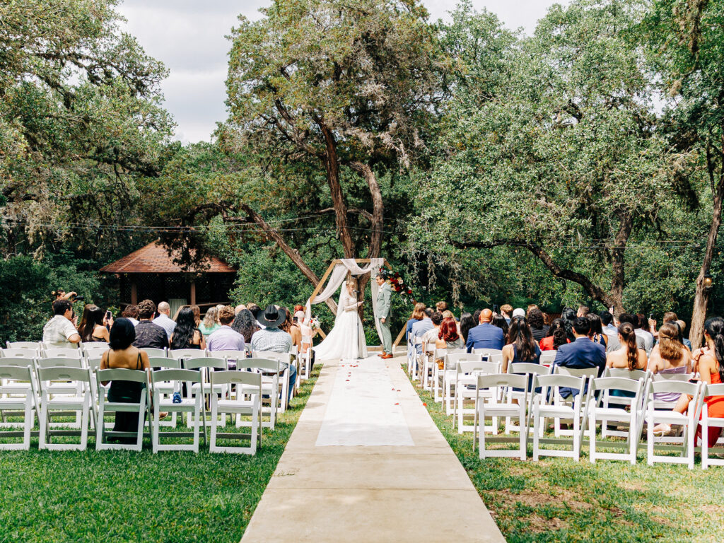 An outdoor wedding ceremony with guests seated on white chairs facing a couple standing under a floral arch. Trees and greenery surround the setting, and a small gazebo is visible in the background.