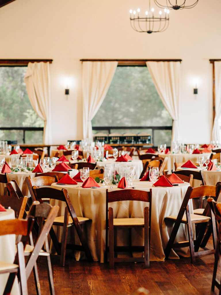 Elegant dining hall with round tables covered in beige tablecloths, set with glassware and red napkins, surrounded by wooden chairs. Large windows with white curtains let in natural light.