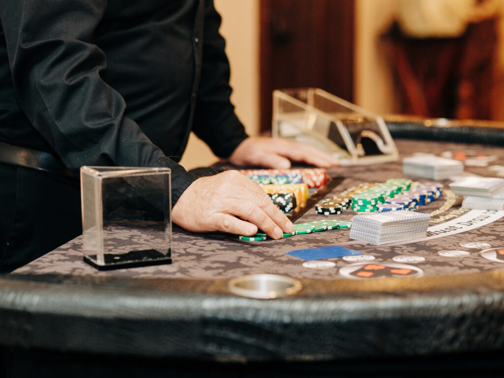 A person in a black shirt stands behind a casino table, organizing colorful poker chips and playing cards. The table has stacks of chips, a card shoe, and a discard tray.