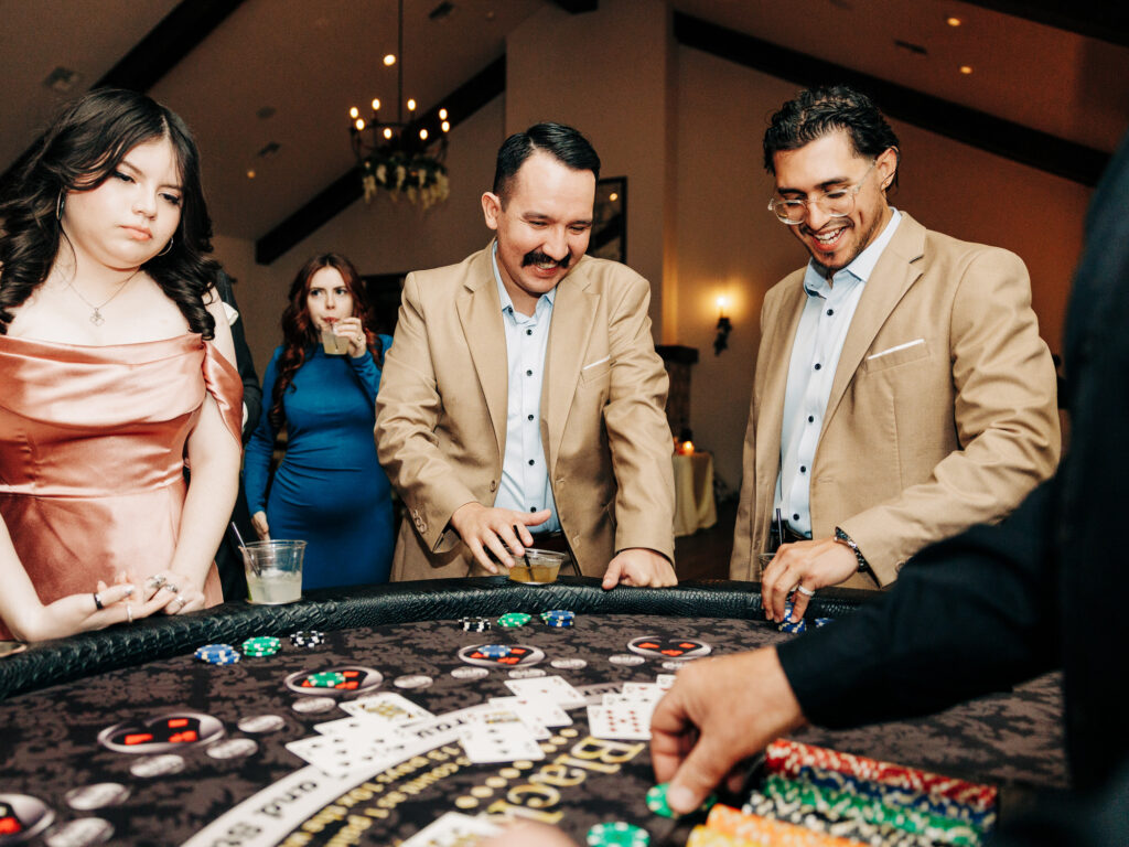 Four people stand around a blackjack table indoors, two men in tan blazers smiling as they play, while a woman in a pink dress looks on with a neutral expression. Chips and cards are visible on the table.