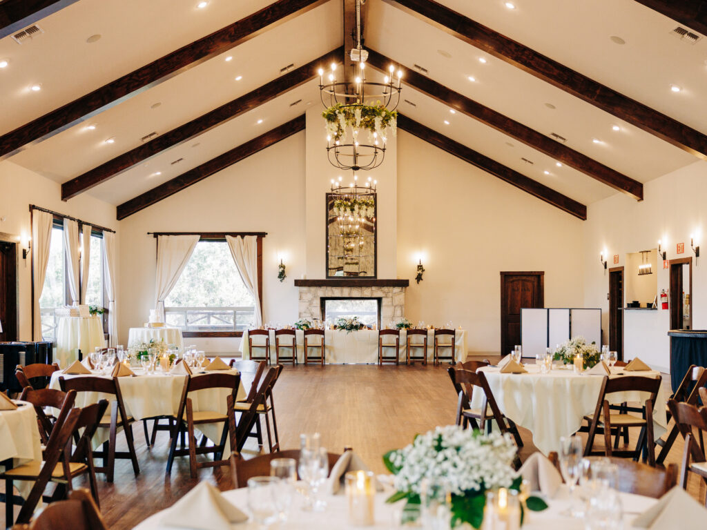 A spacious, elegant banquet hall with wooden beams, round tables set for a formal event, white tablecloths, floral centerpieces, and a decorative chandelier. Large windows and natural light fill the room.
