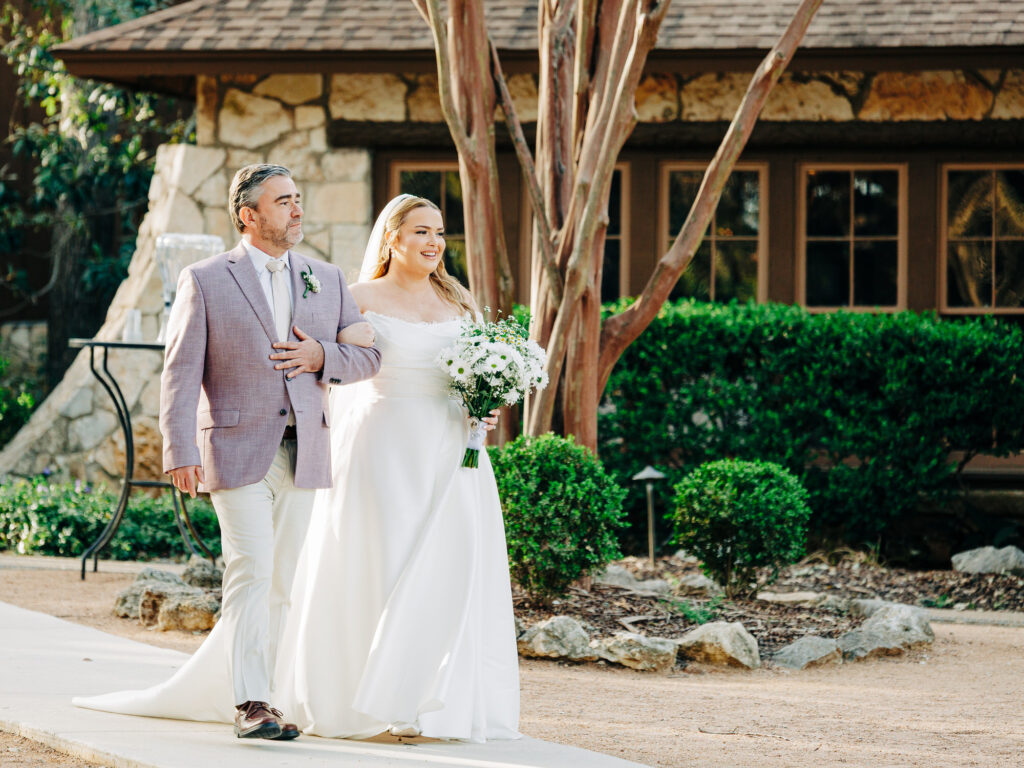 A bride in a white gown holds a bouquet and walks arm-in-arm with a man in a light jacket and pants outdoors, with greenery and a rustic building in the background.