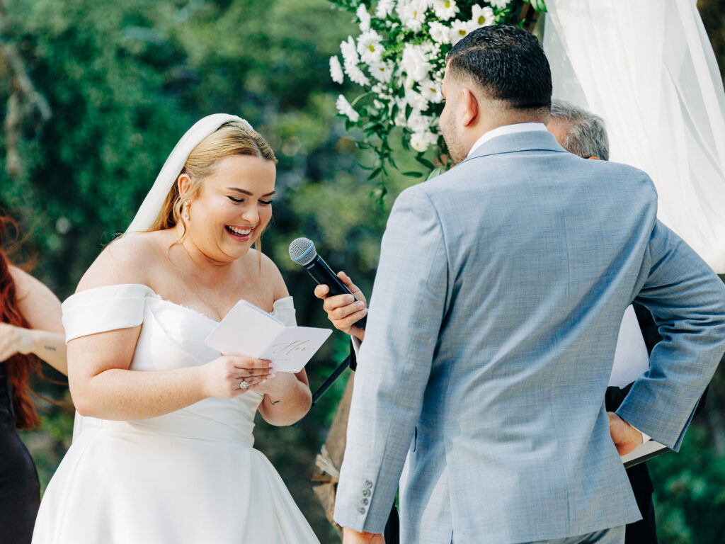 A bride in a white dress smiles while reading vows from a booklet, holding a microphone. The groom, in a light blue suit, stands facing her during an outdoor wedding ceremony with greenery and white flowers in the background.