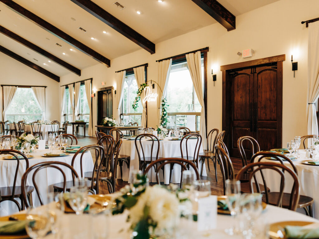 A bright, elegant banquet hall with round tables set for an event, featuring white tablecloths, wooden chairs, gold accents, floral centerpieces, and large windows with sheer curtains.