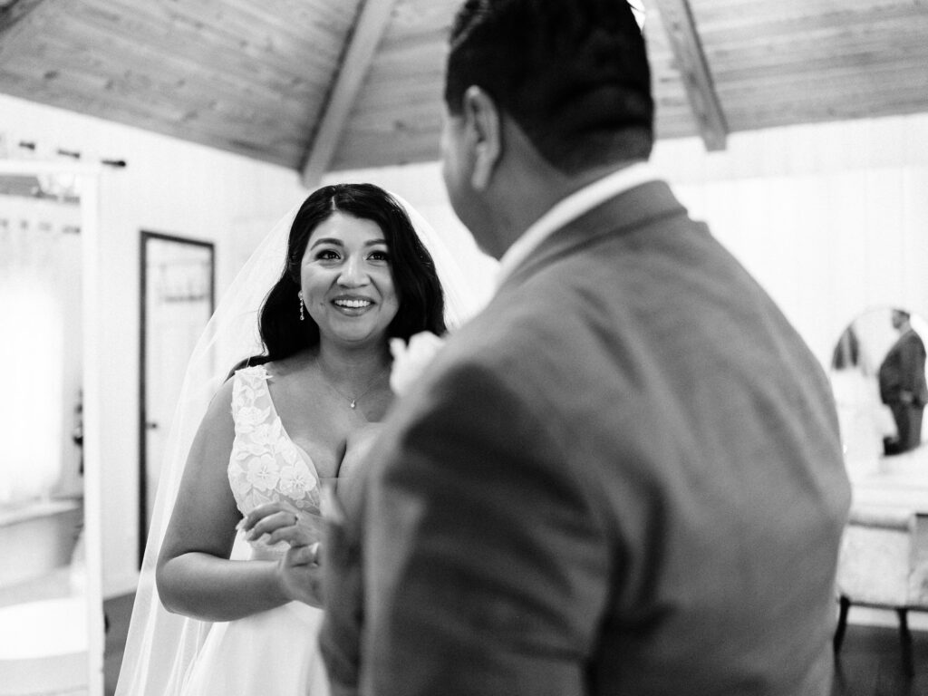 A bride in a white gown and veil smiles warmly at a man in a suit inside a bright, rustic room with wooden ceilings. The scene captures a joyful, candid moment before a wedding ceremony.