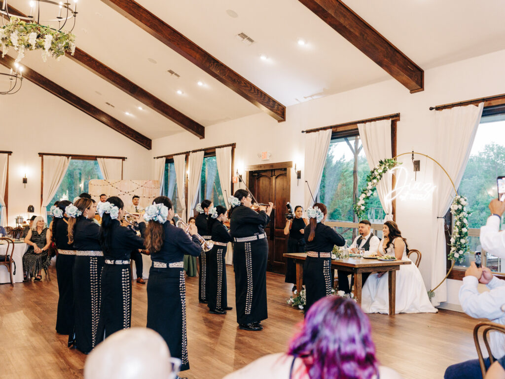 A mariachi band performs indoors at a wedding reception for a bride and groom seated at a decorated table, surrounded by guests in a bright, elegant venue with wooden beams and large windows.