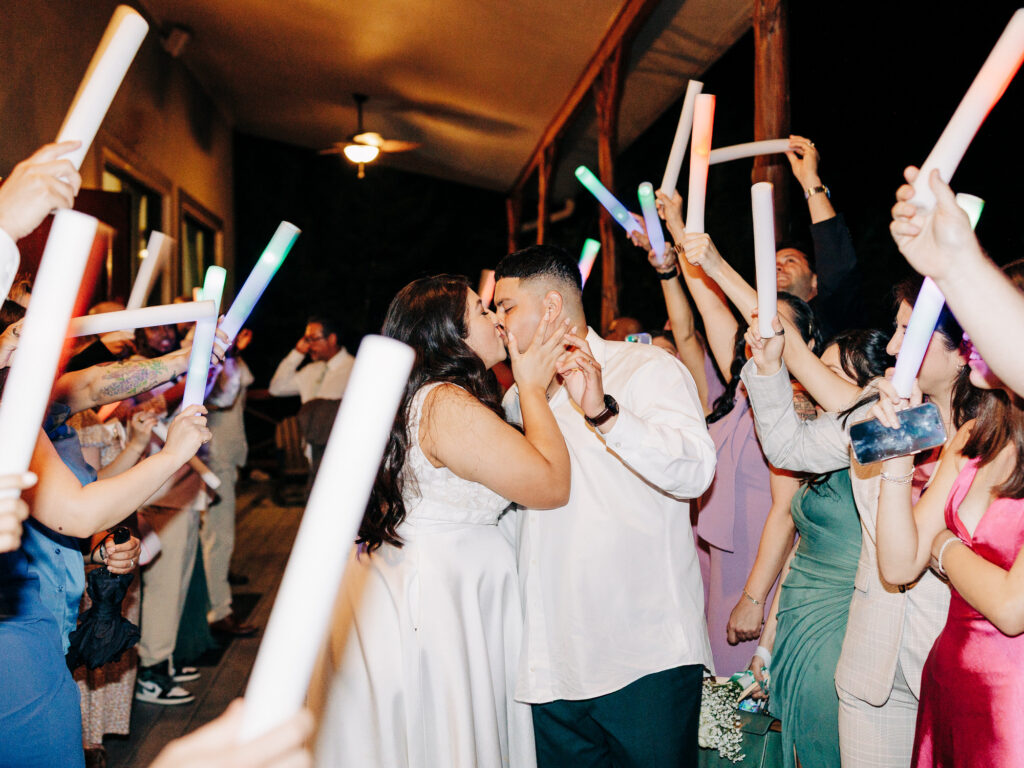 A bride and groom kiss while wedding guests surround them, holding up glowing neon sticks in celebration on a porch at night. The couple stands in the center, dressed in white, with joyful guests on either side.