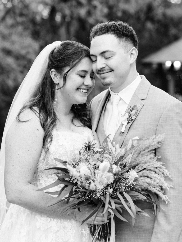 A bride and groom stand close together outdoors, smiling with their eyes closed. The bride holds a bouquet of flowers and wears a lace dress with a veil; the groom is in a light suit and tie. The photo is in black and white.
