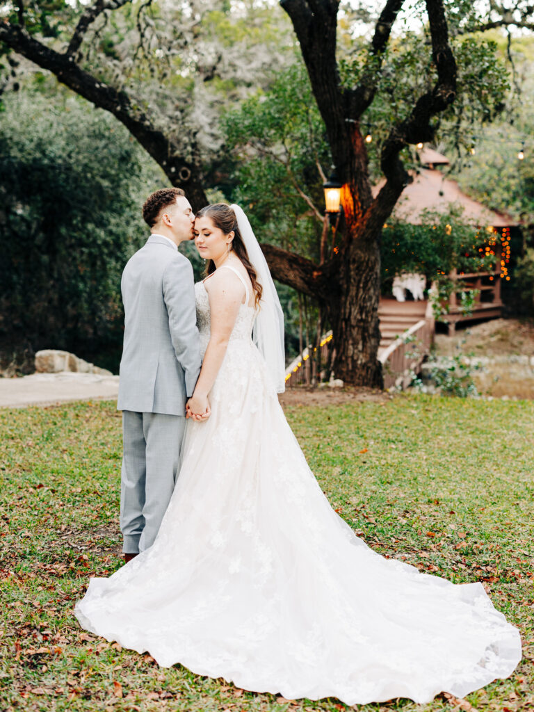 A bride in a long white gown and veil holds hands with a groom in a light gray suit, standing outdoors on grass with trees and a rustic, lit gazebo in the background.