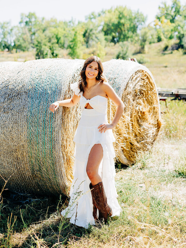 A young woman in a white dress with a high slit and brown cowboy boots stands smiling in front of large round hay bales in a sunny field with green trees in the background.
