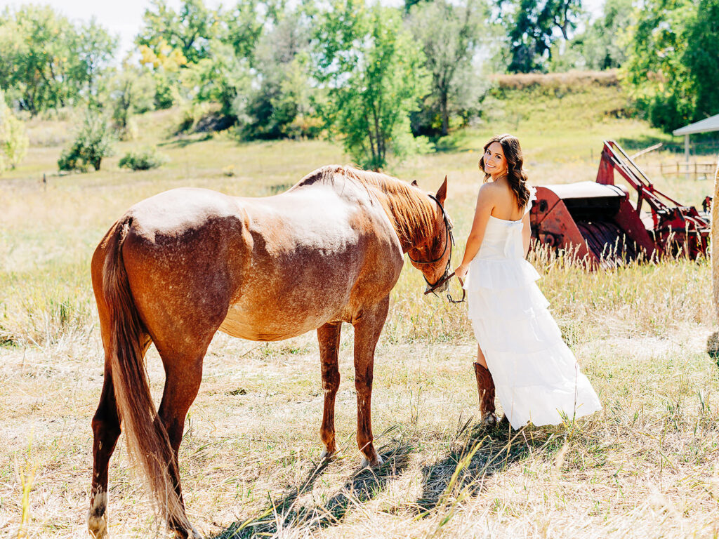A woman in a white dress and brown boots stands in a sunny field, holding the reins of a brown horse. She looks back and smiles, with green trees and a red tractor in the background.