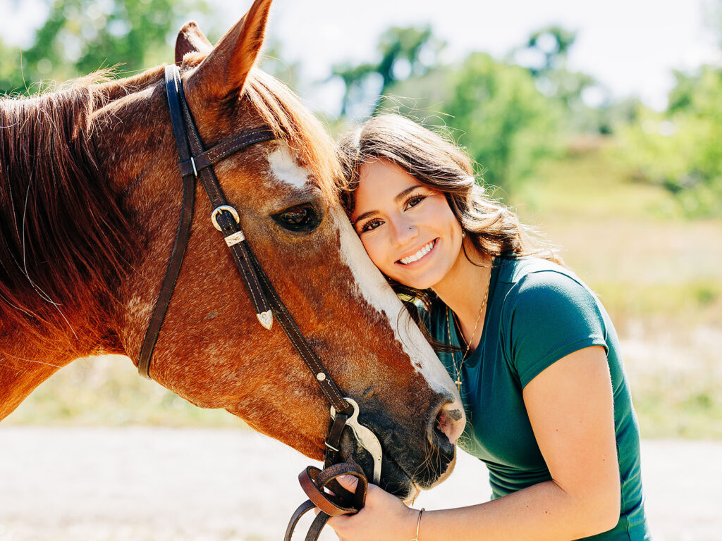 A young woman with wavy brown hair smiles as she leans her head affectionately against a brown horse’s face, holding its bridle, outdoors on a sunny day with green trees in the background.