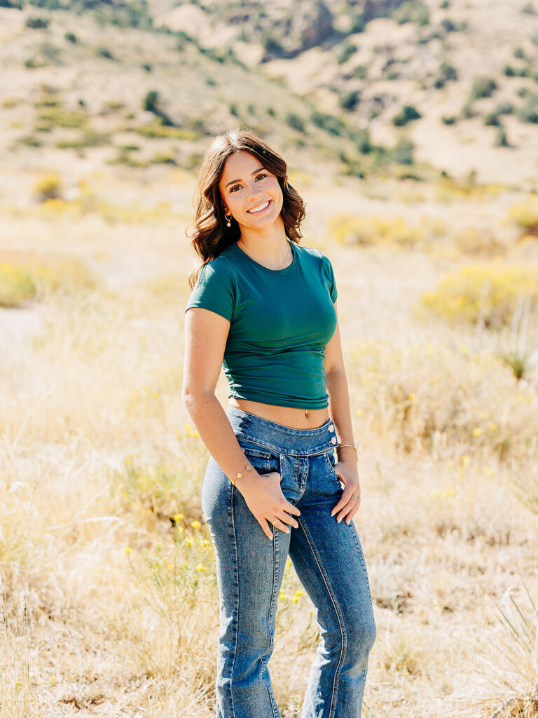 A young woman with long brown hair stands outdoors in a dry, grassy field with hills in the background. She is wearing a green fitted shirt and blue jeans, smiling and looking at the camera.