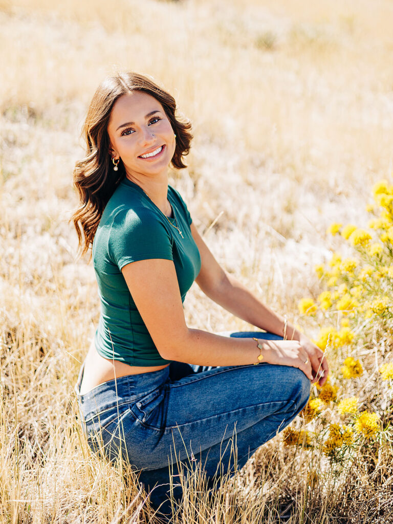 A young woman with wavy brown hair, wearing a green top and jeans, kneels and smiles in a sunlit, grassy field with yellow wildflowers.