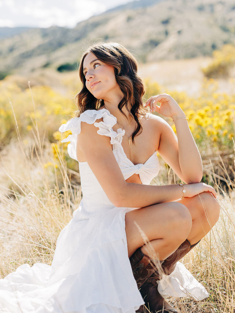 A woman in a white dress and brown boots poses in a sunny field with yellow wildflowers, mountains visible in the background. She crouches, looking thoughtfully to the side, with one hand raised to her hair.