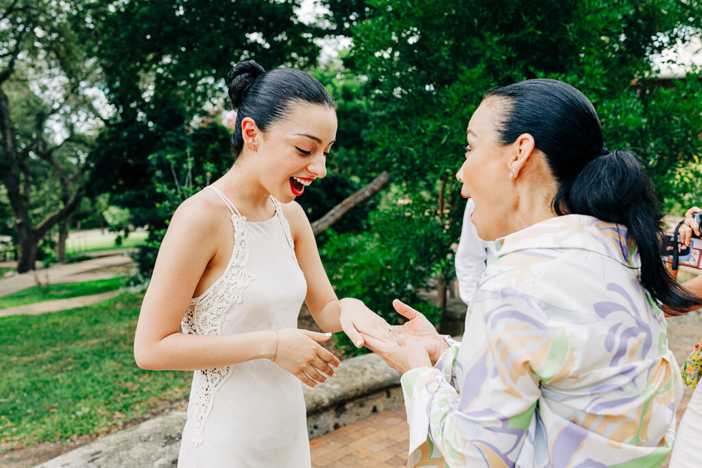 Two women stand outdoors in a park-like setting at the McNay Art Museum, one in a white dress joyfully showing her hand after a proposal, while the other looks excited and surprised amid lush greenery and trees.