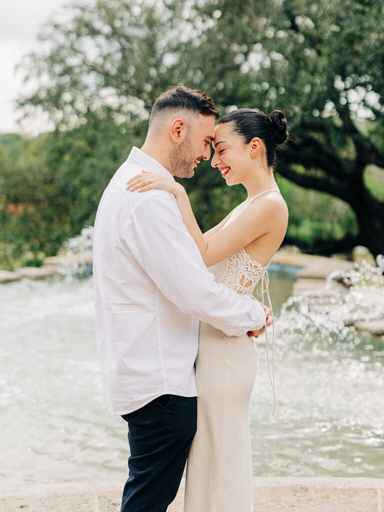 A couple stands close together, smiling and touching foreheads by a fountain outdoors at the McNay Art Museum. The woman wears a white dress, and the man wears a white shirt and dark pants&mdash;capturing the magic of a proposal. Trees and water are in the background.