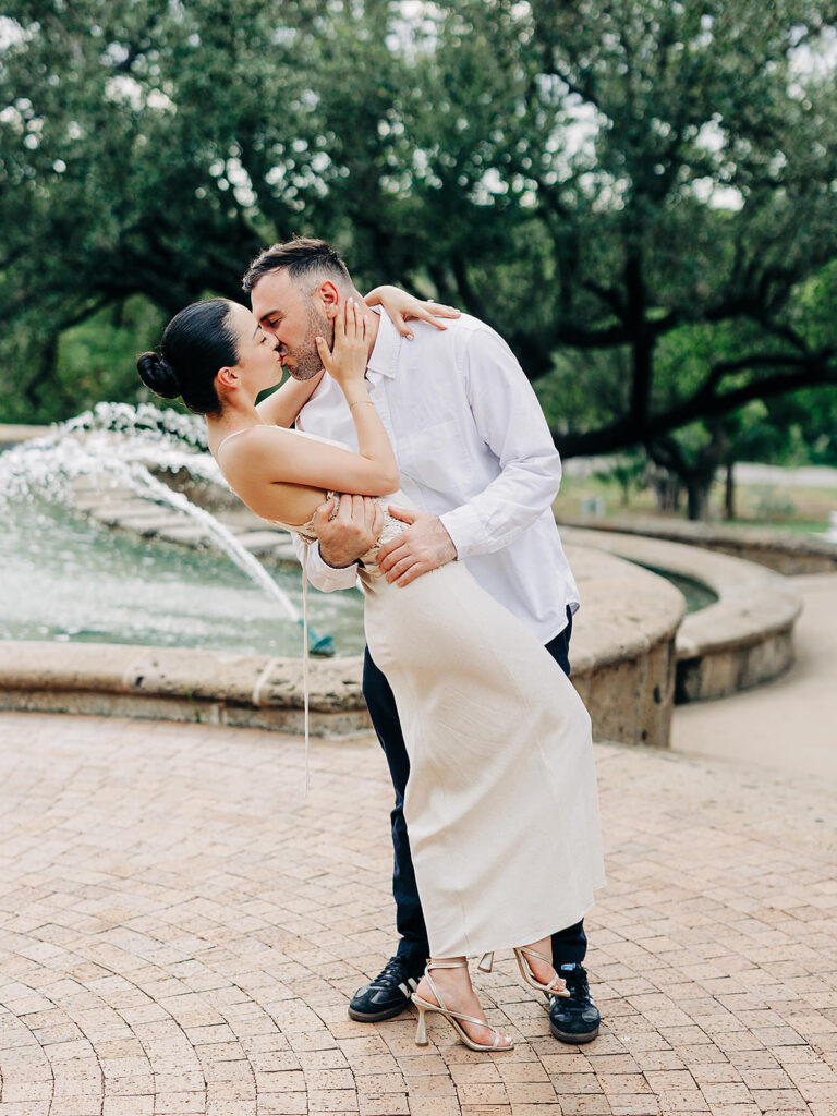 A couple dressed in white kisses passionately as the man dips the woman beside a fountain at the McNay Art Museum, surrounded by trees and a stone pathway&mdash;capturing a romantic proposal in an outdoor park setting.