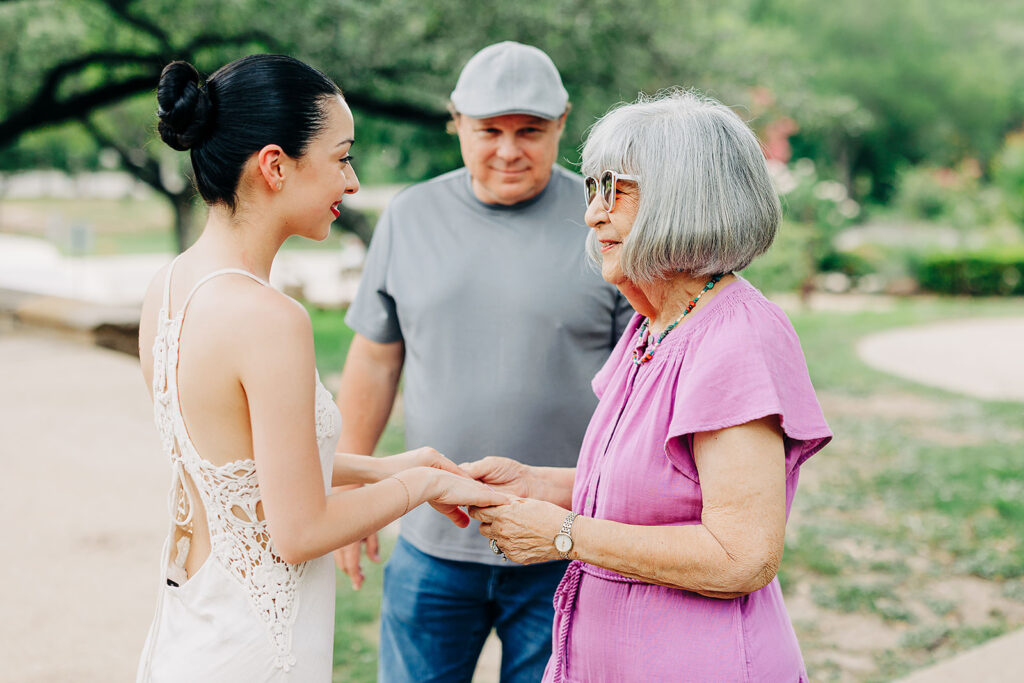 A young woman in a white dress holds hands and smiles with an older woman in a purple outfit outdoors at the McNay Art Museum, while an older man in a gray shirt and cap stands behind them, also smiling.