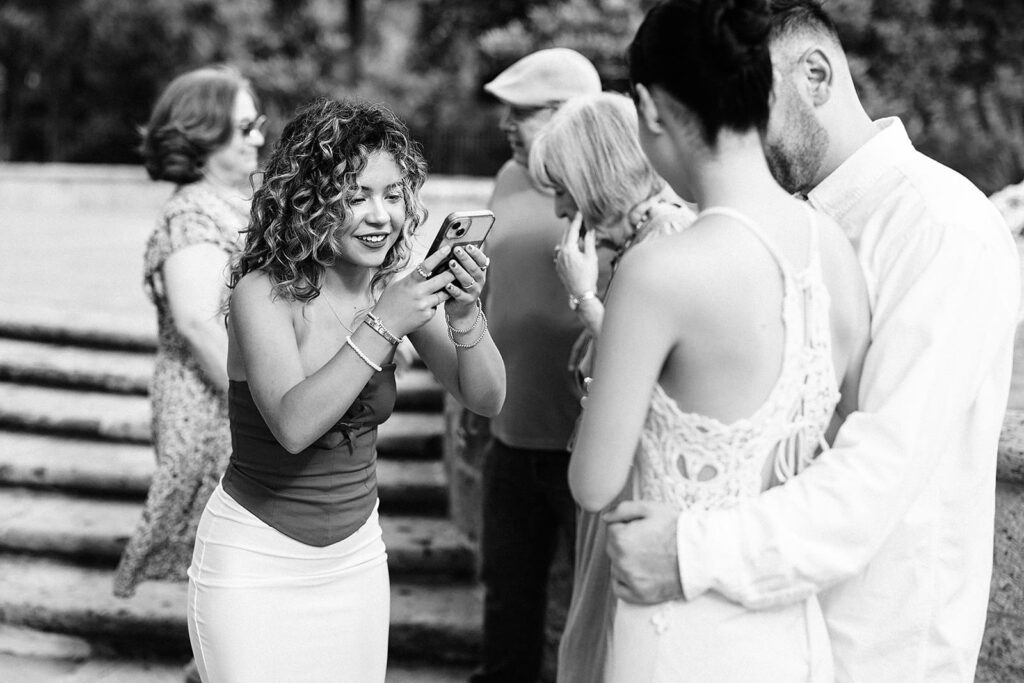 A woman smiles and takes a photo with her phone of a couple, one in a wedding dress, at the McNay Art Museum, while others stand in the background outdoors. The scene appears joyful and candid.