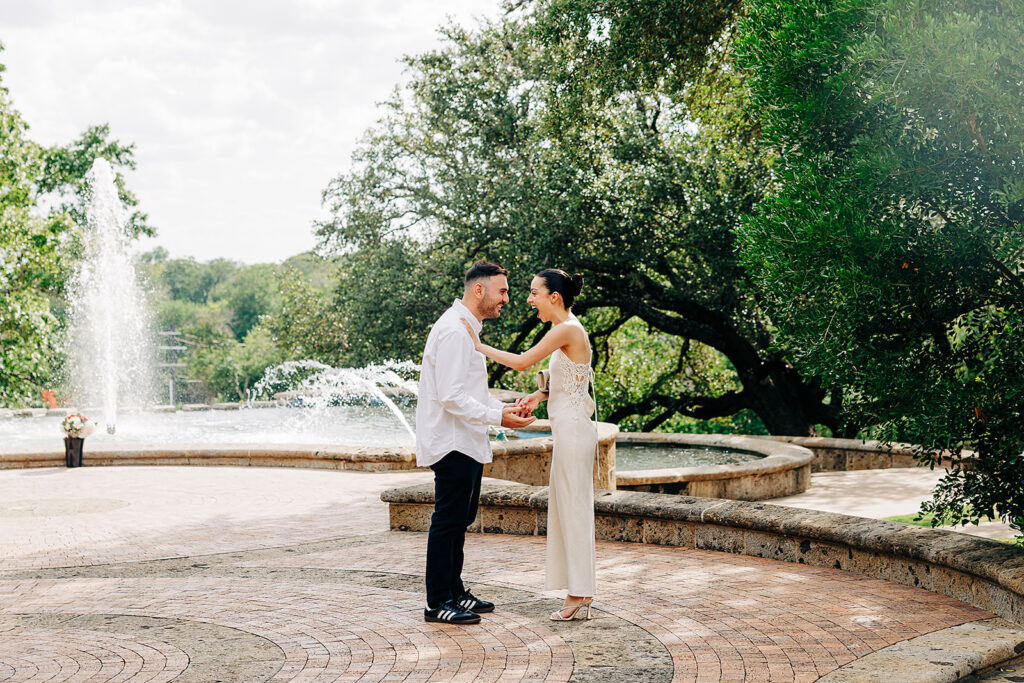 A couple stands facing each other, holding hands in front of a large fountain at the McNay Art Museum, surrounded by trees. The woman is smiling and dressed in white, while the man wears a white shirt and black pants&mdash;perfect for a proposal moment.