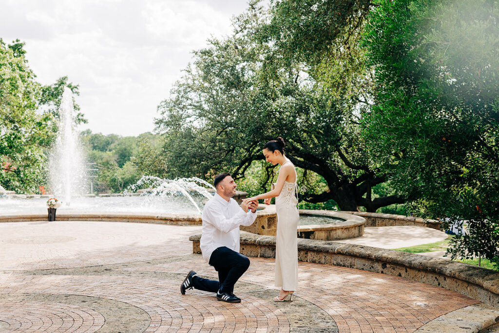 A man kneels on one knee, making a heartfelt proposal to a woman in a white dress. They are outdoors at the McNay Art Museum, surrounded by a circular brick patio, stone fountain, and tall green trees in the background.