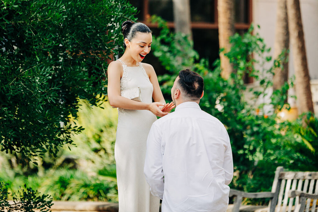 A man kneels in a heartfelt proposal to a surprised, smiling woman in a white dress outdoors at the McNay Art Museum, surrounded by lush greenery.