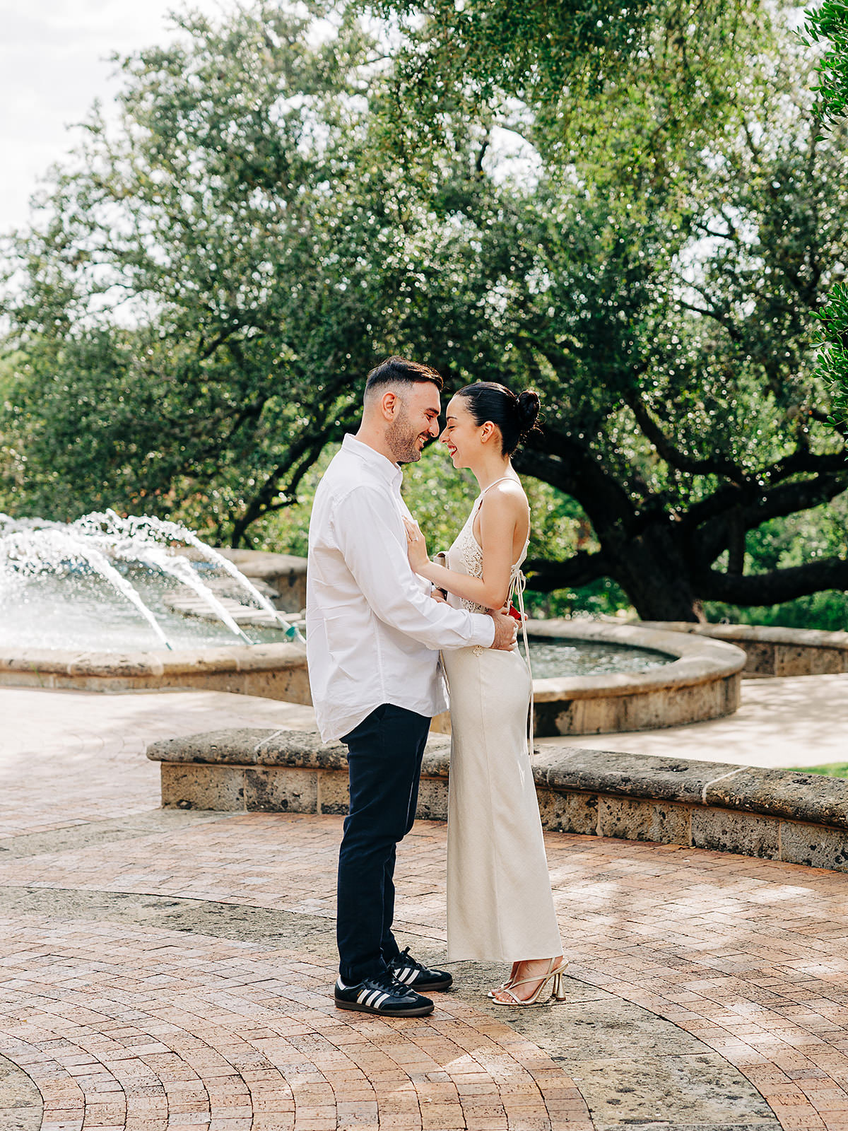 A couple stands close together, smiling and holding each other in front of a stone fountain with water jets at the McNay Art Museum, surrounded by lush green trees—a perfect park setting for a romantic proposal.