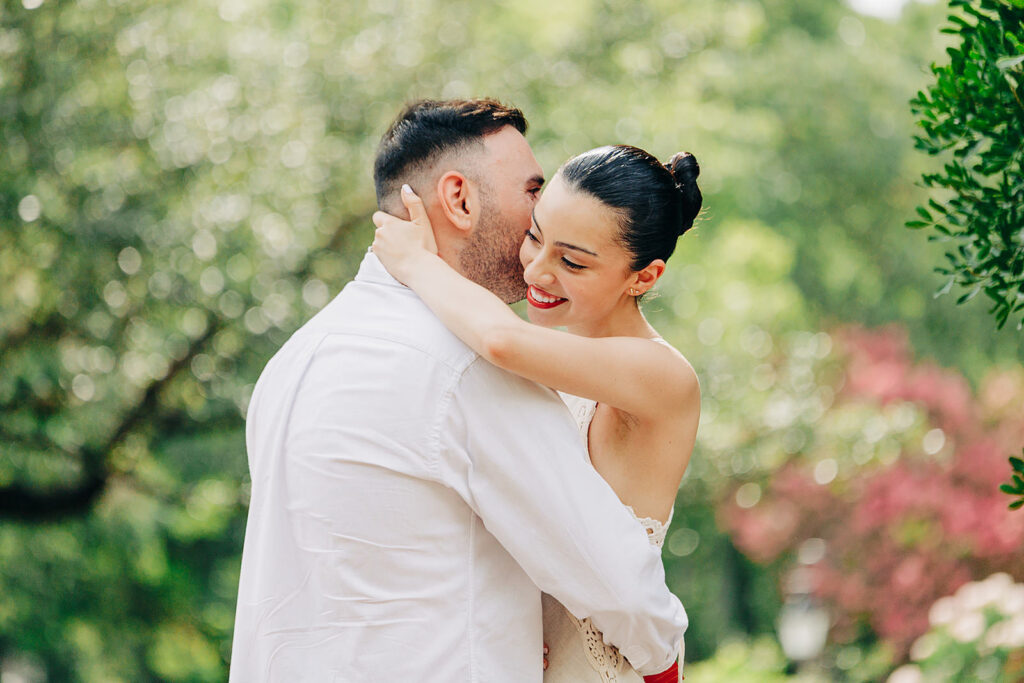 A couple embraces outdoors at the McNay Art Museum; the woman, smiling, wraps her arms around the man&rsquo;s neck as he kisses her cheek&mdash;capturing a joyful proposal amid lush green trees and blurred colorful foliage.