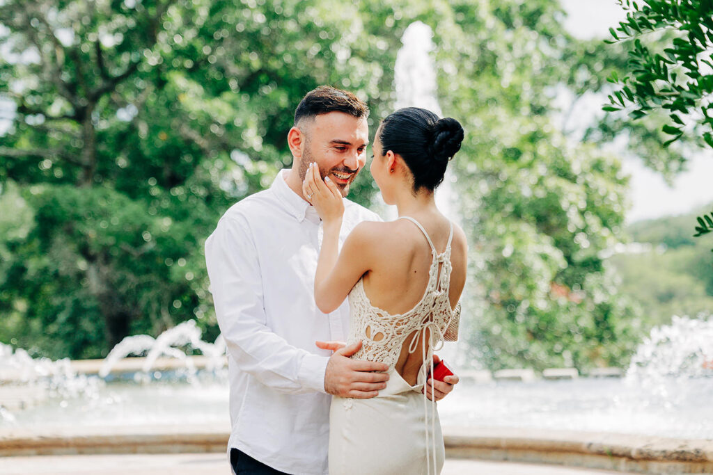 A couple stands in front of a fountain at the McNay Art Museum; the woman touches the man's face affectionately while he smiles. They appear happy and dressed up, with trees and water jets in the background&mdash;perhaps after a proposal.
