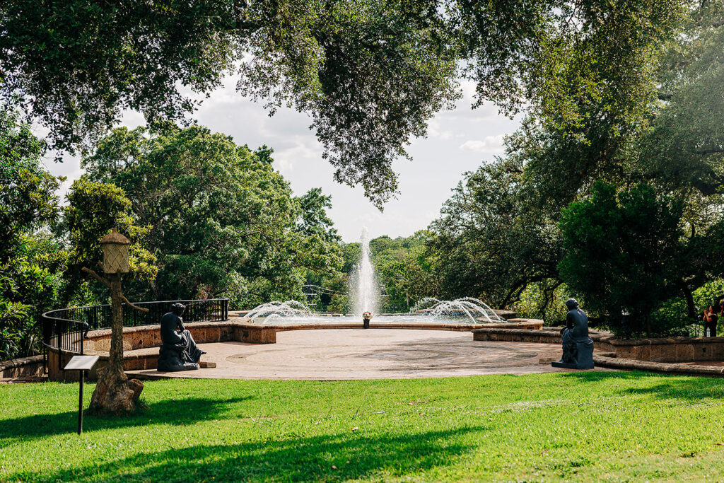 A circular fountain sprays water upward at the McNay Art Museum, surrounded by trees and greenery. Two statues sit on either side, making it a picturesque spot for a proposal as sunlight filters through the leaves onto the grassy area.