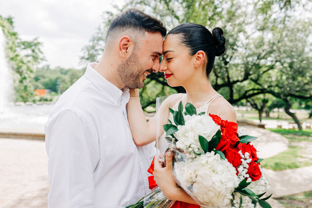 A couple stands close together outdoors at the McNay Art Museum, touching foreheads and smiling. The woman holds a bouquet of red and white flowers, perhaps fresh from a proposal, with trees and a fountain visible in the background.