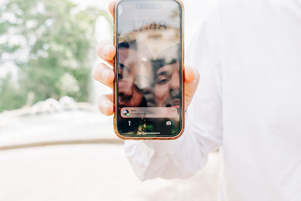 A person in a white shirt holds a smartphone close to the camera, showing the lock screen with a reflection of their smiling face. The background is outdoors near the McNay Art Museum, with trees and a blurred fountain, hinting at a special proposal.