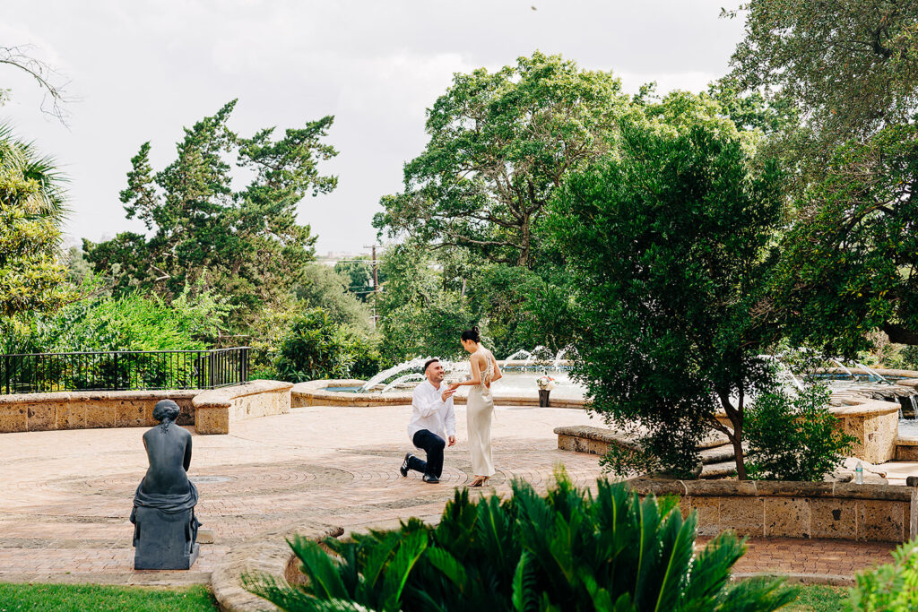 A man kneels to propose to a woman in a sunny, tree-lined park with stone pathways at the McNay Art Museum. A bronze statue sits nearby, and a fountain can be seen in the background as the couple shares their special proposal moment.