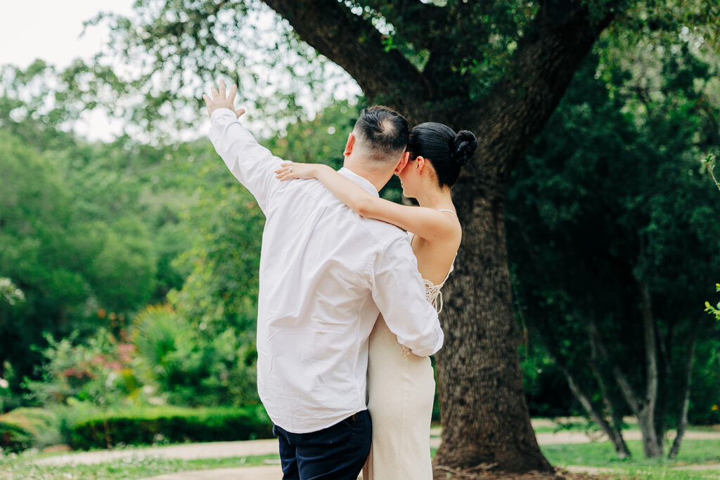 A couple stands outdoors at the McNay Art Museum with their backs to the camera. The woman in a cream dress and the man in a white shirt embrace, as he raises one arm as if waving, surrounded by lush greenery&mdash;a romantic proposal moment.