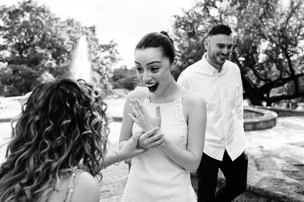 A woman in a white dress looks surprised and excited, showing her hand to a friend after a proposal, as a man stands smiling behind them in an outdoor park setting at the McNay Art Museum.