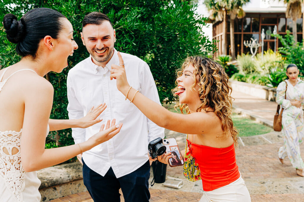 Three people stand outside, laughing and smiling together. One woman gestures playfully toward another, who laughs with her mouth open. A man in a white shirt smiles in the middle&mdash;perhaps celebrating a proposal at the scenic McNay Art Museum.