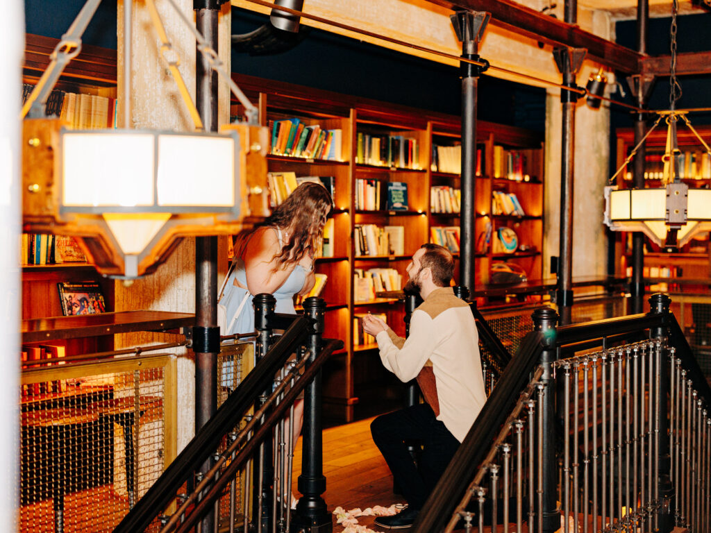 A man kneels in front of a woman on a balcony in a warmly lit library, proposing to her&mdash;one of the most enchanting San Antonio proposal locations. Bookshelves filled with colorful books and hanging lights illuminate the heartfelt scene.