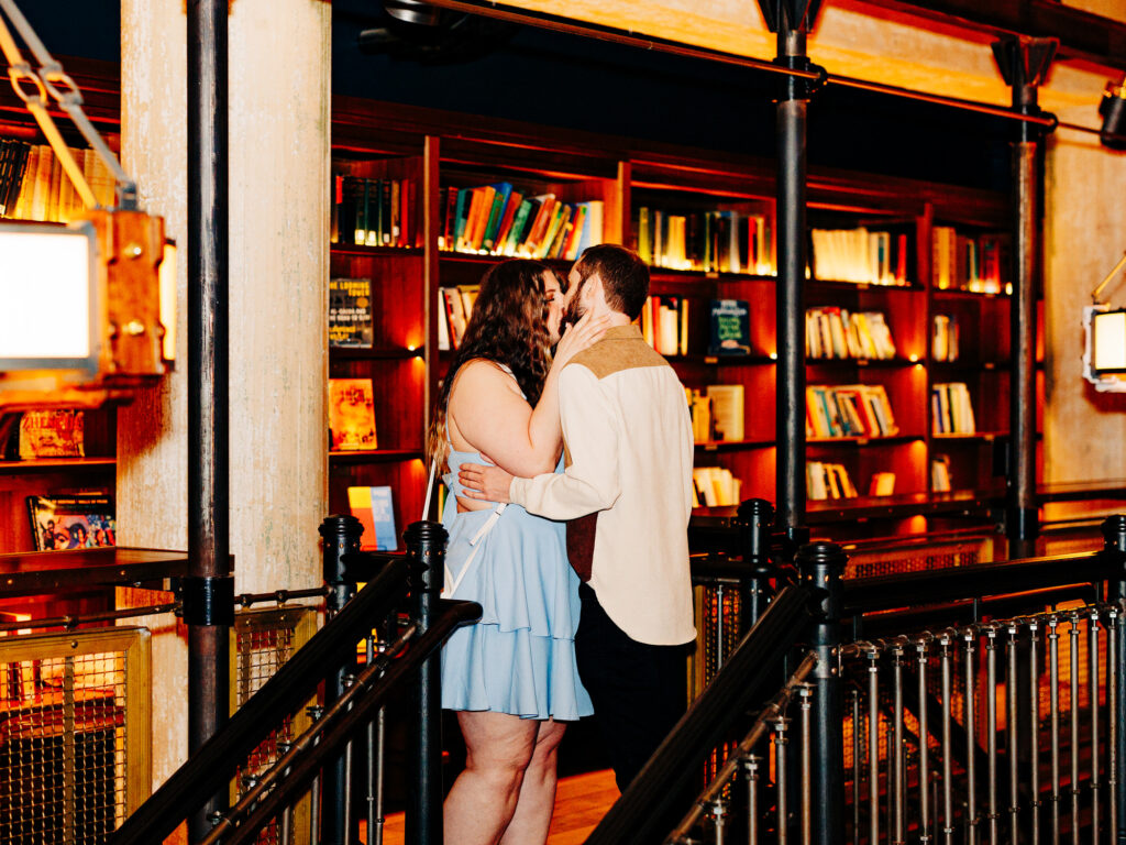 A couple embraces and kisses in front of a bookshelf filled with books in a warmly lit library, standing near a staircase with black railings&mdash;one of the most romantic San Antonio proposal locations.