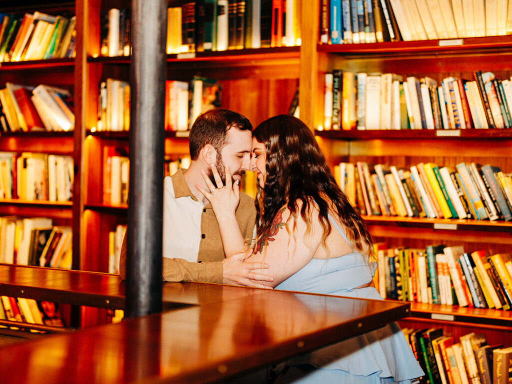 A couple sits closely together at a wooden table in a cozy library, touching foreheads and smiling affectionately&mdash;capturing the charm of unique San Antonio proposal locations. Bookshelves filled with books are in the warmly lit background.