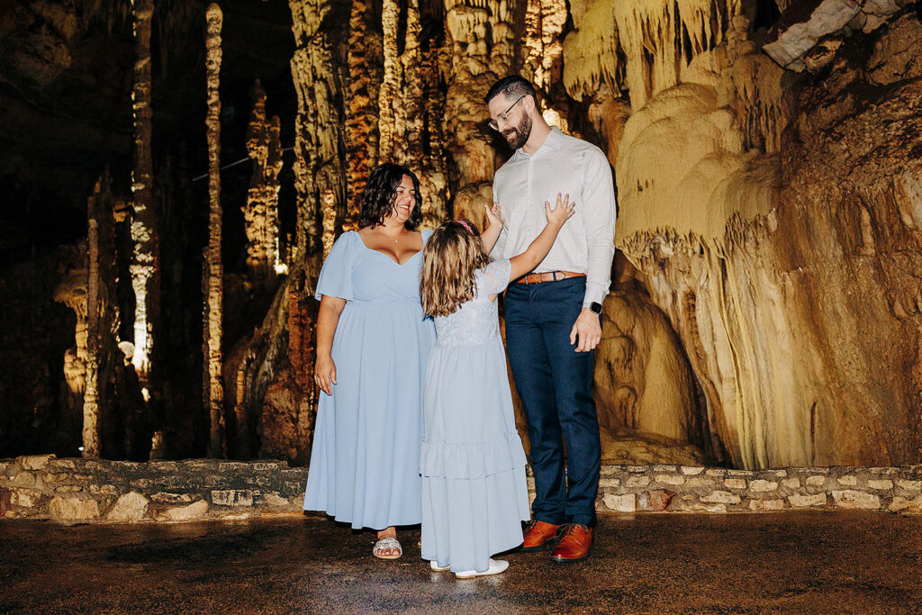 A woman, a man, and a young girl stand together inside Natural Bridge Caverns with dramatic stalactites and rock formations. The girl reaches up towards the man, as if witnessing a touching proposal; all three wear light blue outfits.
