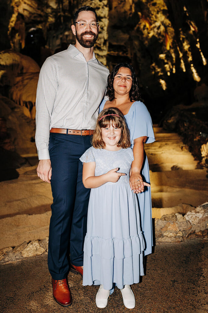 A man, woman, and young girl stand together smiling in the dimly lit Natural Bridge Caverns. The man wears a light shirt and dark pants; the woman and girl wear light blue dresses. The girl stands in front, pointing at something out of frame—perhaps a proposal spot.