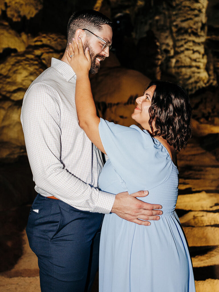 A woman in a light blue dress lovingly embraces a man in glasses and a button-up shirt. They smile at each other while standing close together inside the Natural Bridge Caverns, rocky walls framing their intimate proposal moment.