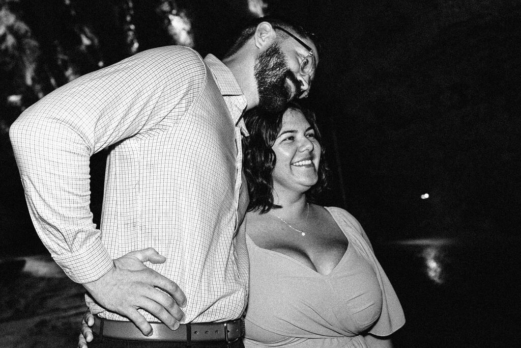 A man and woman stand closely together, smiling and looking towards the right. The image is black and white, capturing a joyful proposal moment at Natural Bridge Caverns, with the man’s arm around the woman’s shoulder.