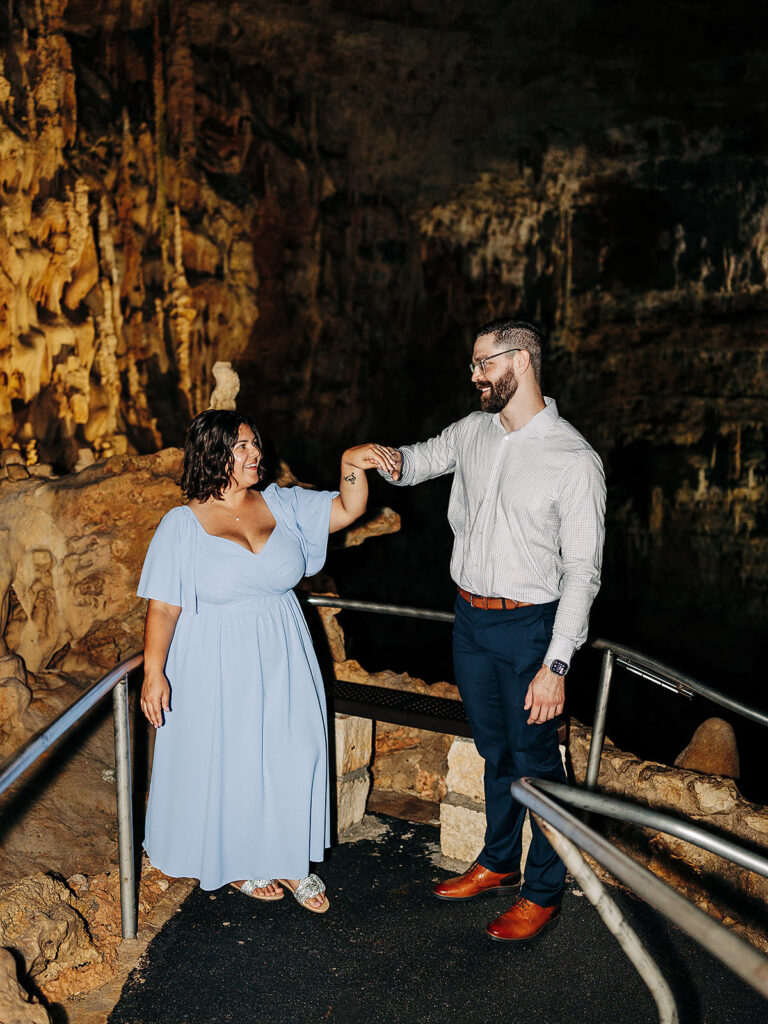 A woman in a light blue dress and a man in a light shirt and dark pants stand together on a cave pathway at Natural Bridge Caverns, smiling and holding hands beneath stunning stalactites—capturing the magic of a proposal among breathtaking rock formations.