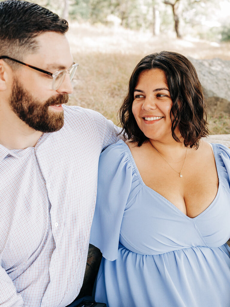 A man with a beard and glasses sits beside a woman in a light blue dress. Outdoors at Natural Bridge Caverns, they look at each other and smile, capturing the joy of their romantic proposal amid trees and grass.