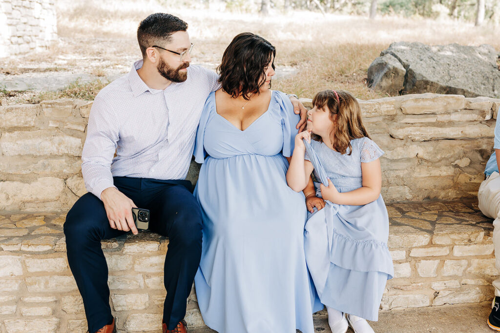 A man, a woman in a light blue dress, and a young girl in a matching dress sit on a stone bench outdoors at Natural Bridge Caverns, smiling and interacting warmly—perhaps celebrating after an unforgettable proposal.