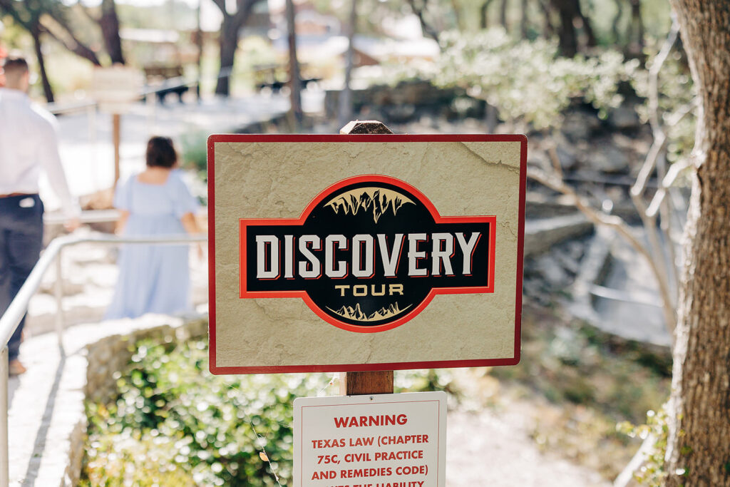 A sign reading Discovery Tour stands on a pathway surrounded by trees at Natural Bridge Caverns. Two people walk away in the background on a sunny day, perhaps planning a proposal. A smaller warning sign is posted below the main sign.
